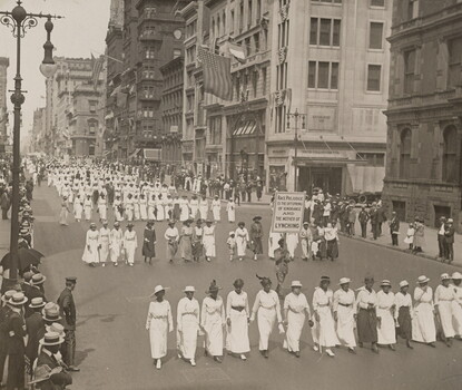 Lines of women primarily in white dresses and hats marching in formation. Between the first and second lines, a man in a dark suit and hat holds a sign reading "RACE PREJUDICE IS THE OFFSPRING OF IGNORANCE AND THE MOTHER OF LYNCHING."