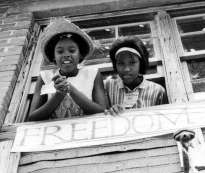 A black-and-white photograph of two Black girls, one wearing a straw hat and the other with a hair band, leaning out of a window above a banner reading "Freedom School." 
