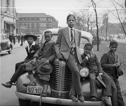 A black-and-white photograph of five Black boys in suits and ties sitting on the hood of a car parked on a tree-lined street in Chicago.