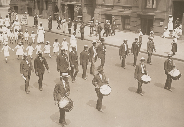 A line of four Black men in dark suits and hats beating small drums, marching ahead of a line of more Black men in dark suits and hats, and girls and women in white dresses (one of whom holds a sign reading "THOUGH SHALT NOT KILL"). People are watching along the sidewalk, including a uniformed police officer holding a nightstick.