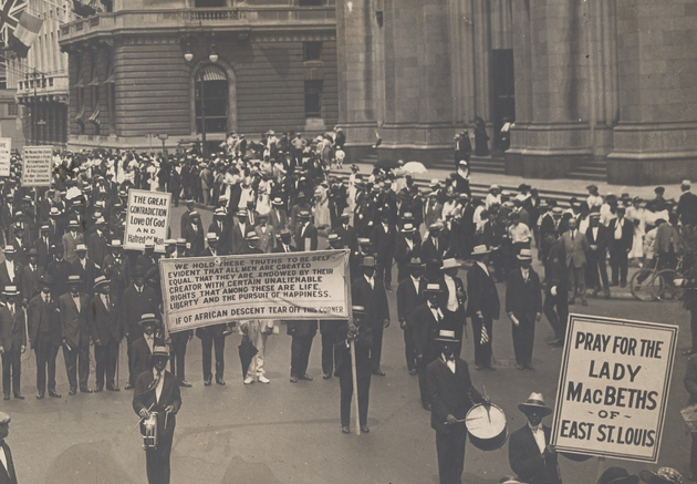 A large group of Black men in dark suits and hats, some holding signs (including one reading "PRAY FOR LADY MACBETHS OF EAST ST. LOUIS" in the right foreground), marching in formation past a gothic church and other buildings on the right side of the photograph.