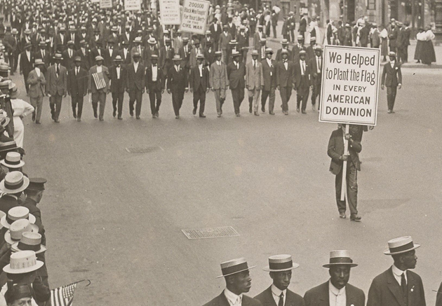 Men in dark suits and hats marching in formation — a single row, then two men holding signs (reading "We Helped to Plant the Flag IN EVERY AMERICAN DOMINION" and "TAXATION WITHOUT REPRESENTATION IS TYRANNY"), then a mass of multiple lines. People are watching along the sidewalk.