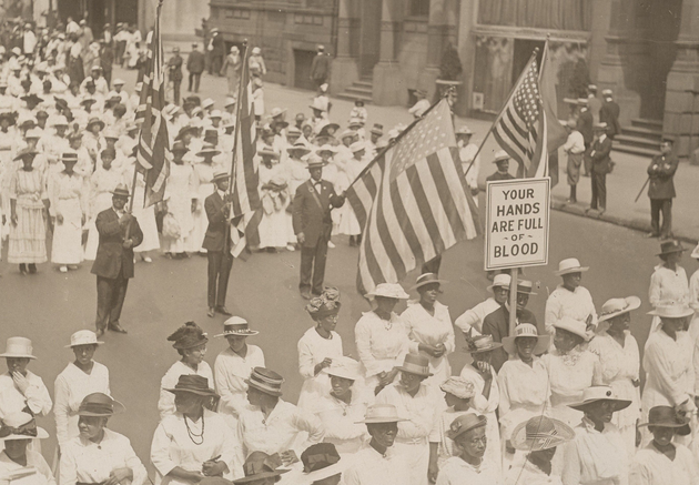 Two large groups of Black women in white dresses and hats (some dark, some white) marching in formation. Between these two groups are four Black men in dark suits, ties, and hats, waving large American flags. In the first group of women, one woman holds up a sign reading, "YOUR HANDS ARE FULL OF BLOOD."