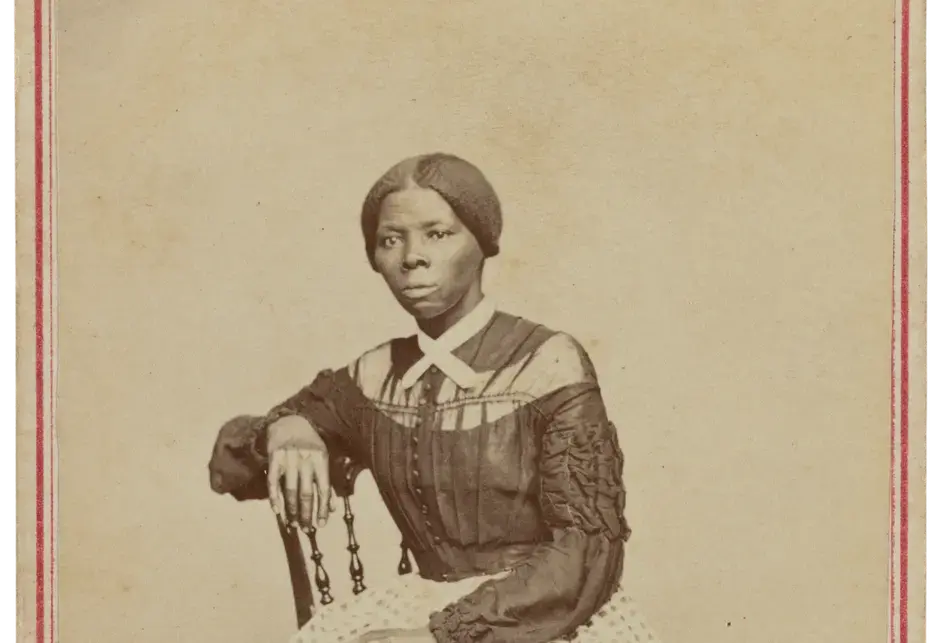 Harriet Tubman, a Black woman, seated in an interior room. She is positioned slightly turned to the right and gazes off camera. Her right hand rests on the back of a wooden chair and her left hand rests in her lap. A boldly patterned carpet covers the floor and the wall or drop behind her is a blank light color. Tubman wears a black bodice that buttons at the center front and has dropped sleeves with heavy ruching and ruffled details on the sleeves.