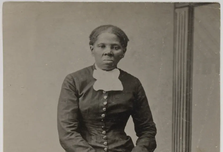 Harriet Tubman, a Black woman, standing in an interior room setting. The full-length portrait shows Tubman facing the photographer in the middle of the frame with her hands resting on the back of an upholstered chair. Tubman wears a dark bodice with metal buttons at the center front and full-length fitted sleeves. A white ruffled lace collar is tied at her neck. Her ankle length skirt is full with gathers at the waist and a wide decorative fabric band around the skirt near the hem.