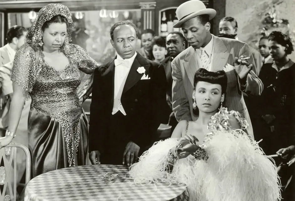 In this film still, a Black woman wearing a dark, sparkly dress, a Black man in a tuxedo with white tie, and a Black man in a light-colored jacket, white shirt, light-colored patterned tie, and light-colored hat with a dark brim stand over a table with a checkered tablecloth, as a Black woman wearing fur sits. 