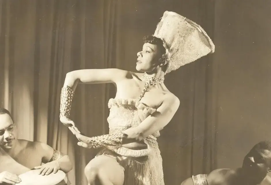Katherine Dunham, a Black woman, dancing in costume (including a large white hat/headdress) with bare feet. In the background, two Black men sit shirtless while drumming.
