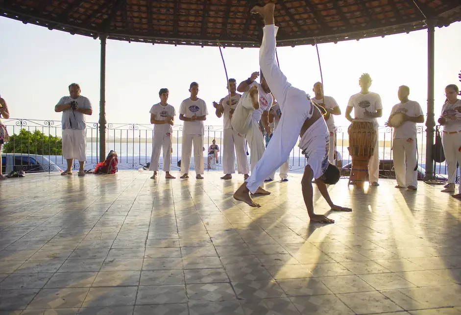 A color photograph of a man wearing a white shirt and pants begins a handstand as a band plays in the background and others look on.