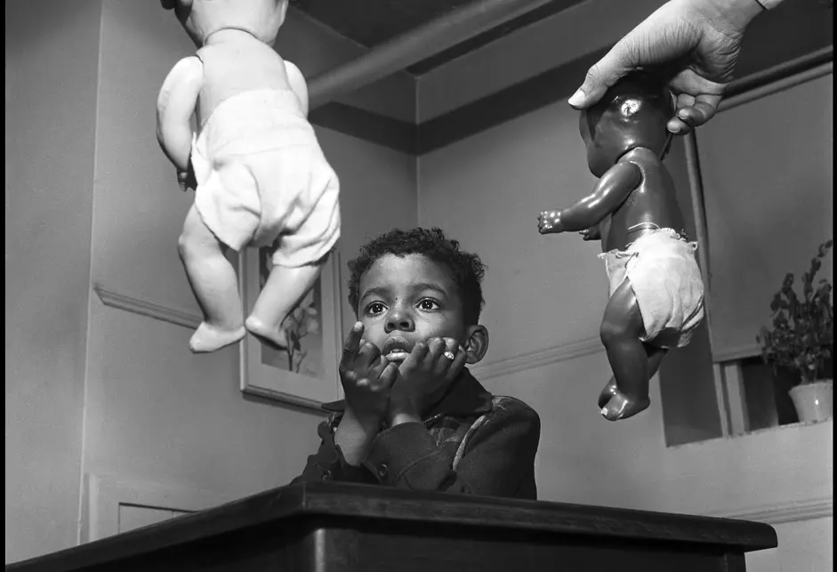 A young Black boy sits, heads in hands, and points toward a white baby doll held in the left hand of Kenneth Clark, a Black man whose white shirt and gray pinstriped jacket sleeves are visible. in Dr. Clark's right hand is a black baby doll.
