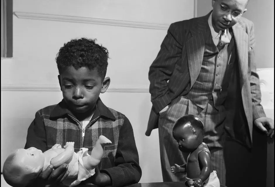 A young Black boy wearing a plaid zip-up sweater holding a white baby doll, as a black baby doll sits upright on a table. Kenneth Clark, a bespectacled Black man wearing a gray pinstriped suit (jacket, vest, pants), white shirt, and patterned tie, looks on in the background.
