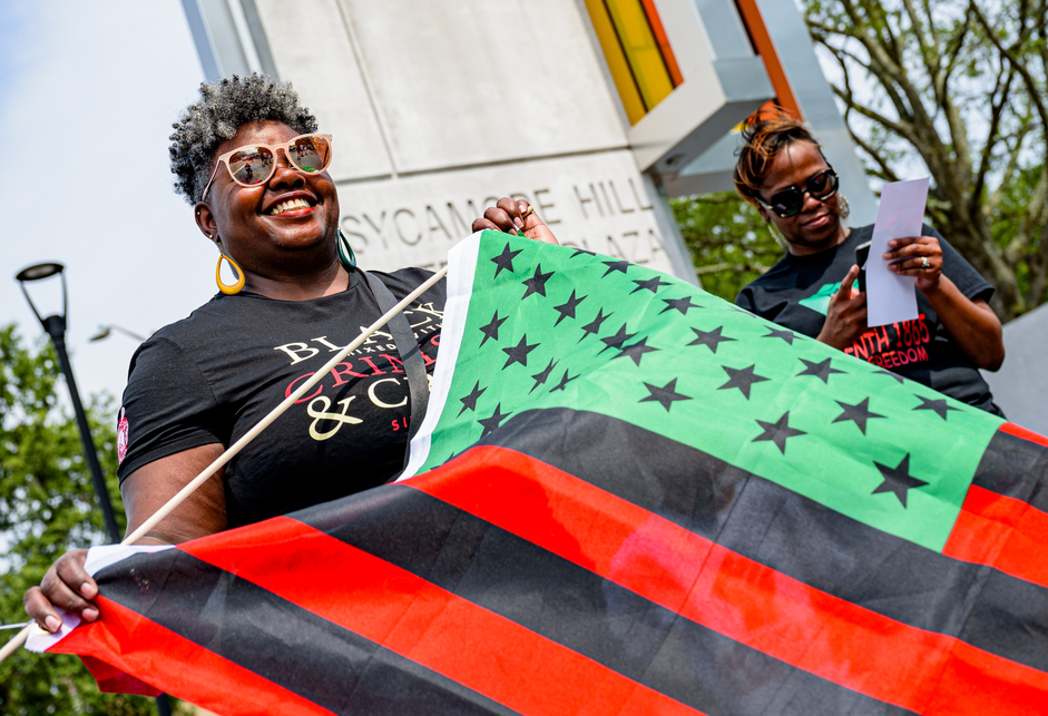 A color photograph of a woman wearing a black t-shirt, yellow hoop earrings, and sunglasses holding an American flag in the colors of the Pan-African flag (red, black, and green).