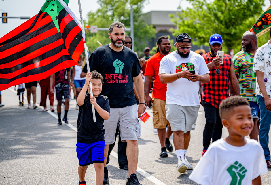 1: A color photograph of a parade in celebration of Juneteenth; men, women, and children predominantly wearing T-shirts and shorts or jeans marching on a city street, with a child holding an American flag in the colors of the Pan-African flag (red, black, and green)  in the left foreground.