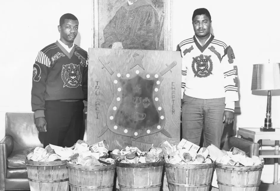 Two young Black men in long-sleeved T-shirts emblazoned the Omega Tau Psi crest across the chest, flanking a large crest for the fraternity, in front of five barrels full of canned and packaged food.