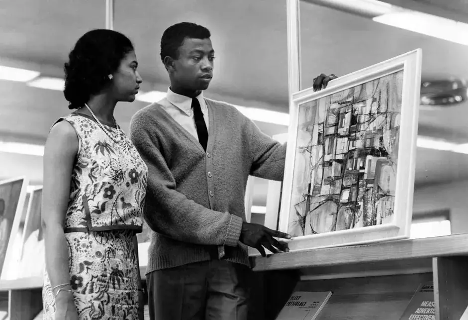 A Black woman wearing a floral dress looking at a painting held by a young Black man wearing a light-colored cardigan sweater, white shirt, and dark tie.