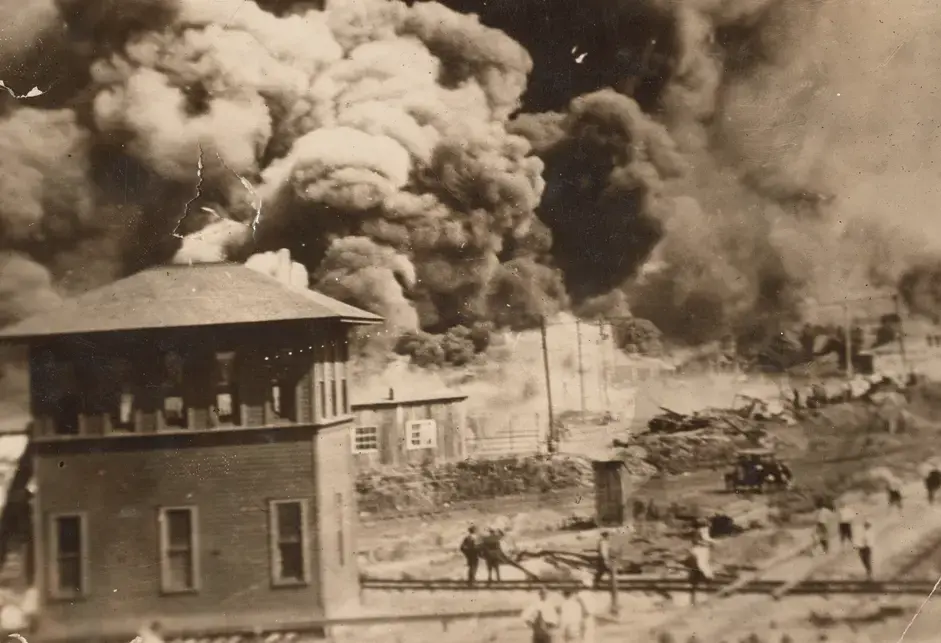 A black-and-white photograph of the Greenwood District of Tulsa, Oklahoma burning during the Tulsa Race Massacre in 1921. The image depicts a building and several people in the foreground watching large plumes of dark smoke rising in the background. The photograph is bent at corners and has loss at top center edge. 