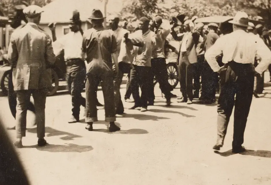 A black-and-white photograph of African American men standing in a loose line, with hands raised in surrender, along what appears to be a residential street. Several White men are visible standing between the line and the camera, many with hands on their hips.