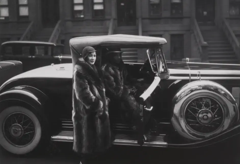 A light-skinned Black woman standing outside a car, and a darker-skinned Black man sitting in the car. Both are wearing heavy fur coats and dark hats.