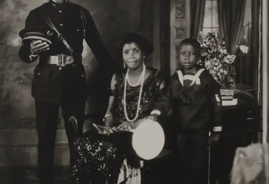 From left to right: A standing Black man in a military-style uniform (dark jacket, light-colored belt and buckle, dark pants, dark shoes, and holding his dark hat; a seated Black woman, wearing a sparkly black dress and shoes; a young Black child wearing a dark sailor suit; and a small dog toy. 