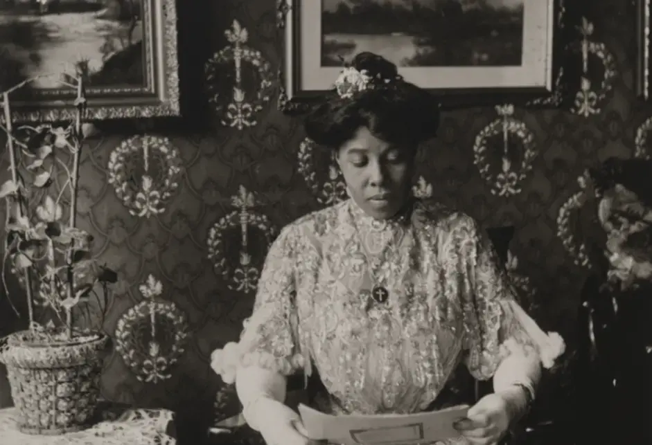 A Black woman in a bejeweled white dress reading in a chair. In the background is a wall with a dark brocade-style wallpaper, adorned with framed images and artwork, and (to the woman's right) a side table with a lace tablecloth and a basket with flowers.