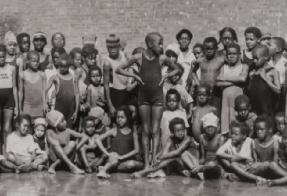 A panoramic image of Black children sitting and standing, with many in bathing suits and swimming caps.