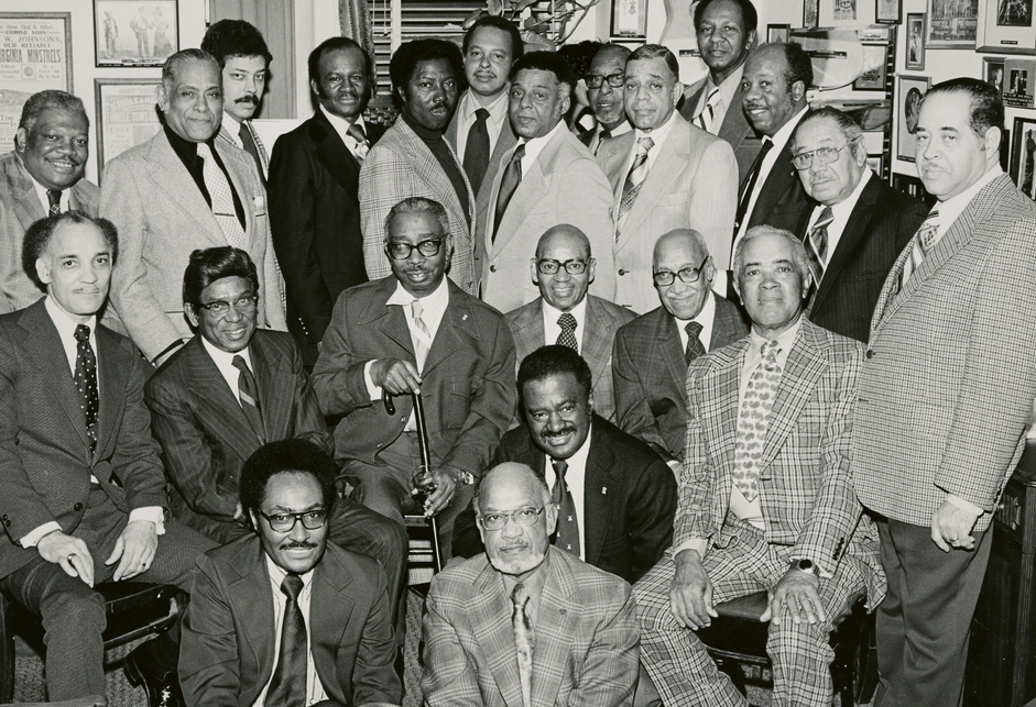  A group of about two dozen Black men, seated and standing, in an office building with many framed photographs and citations hanging on the walls, above a typed and handwritten note.