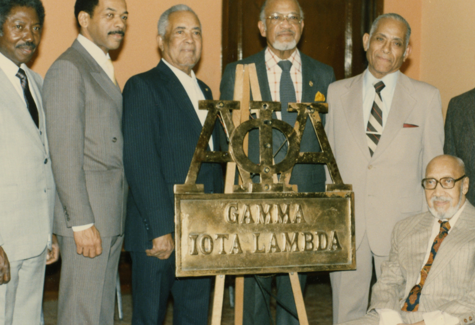 A color photograph of six standing (and one seated) Black men in suits and ties, in front of a metal sign on a wooden easel reading "Alpha Phi Alpha [in Greek letters]: Gamma Iota Lambda."