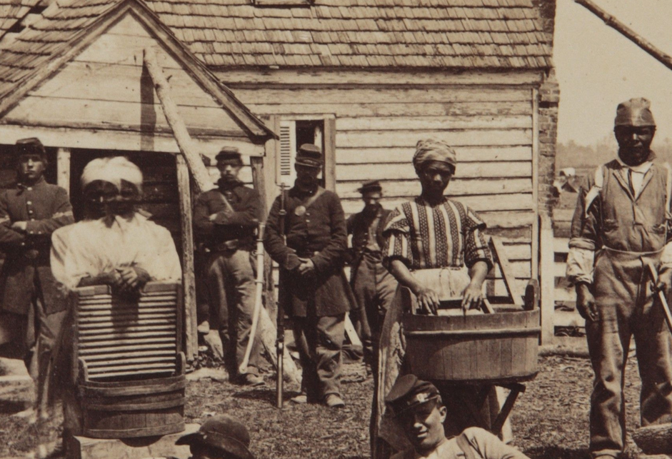 Black men and women laying on the ground and standing (those standing holding tools, such as washboards and basins and an axe) in front of Union soldiers in uniform, themselves in front of a house.
