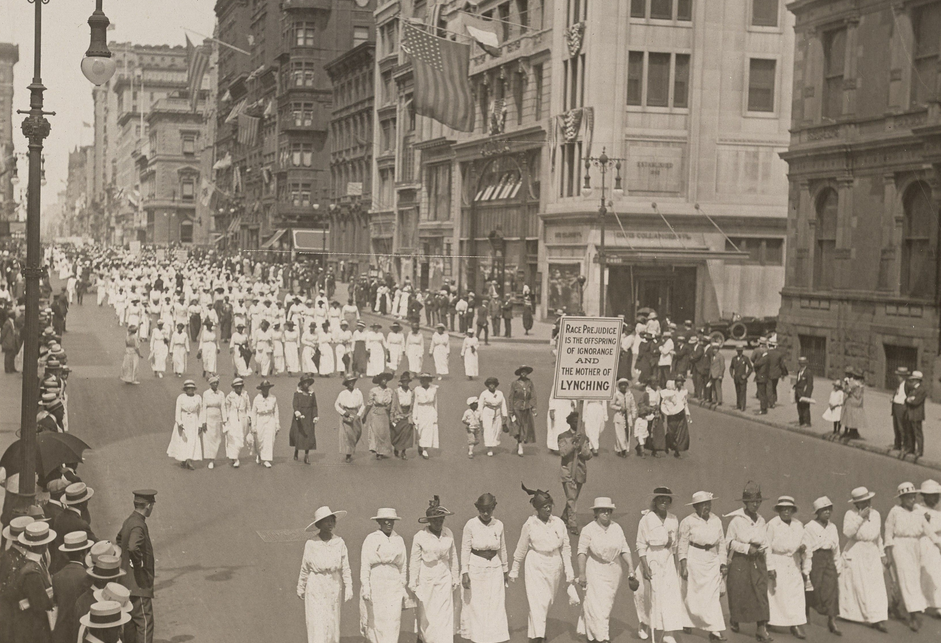 Lines of women primarily in white dresses and hats marching in formation. Between the first and second lines, a man in a dark suit and hat holds a sign reading "RACE PREJUDICE IS THE OFFSPRING OF IGNORANCE AND THE MOTHER OF LYNCHING."
