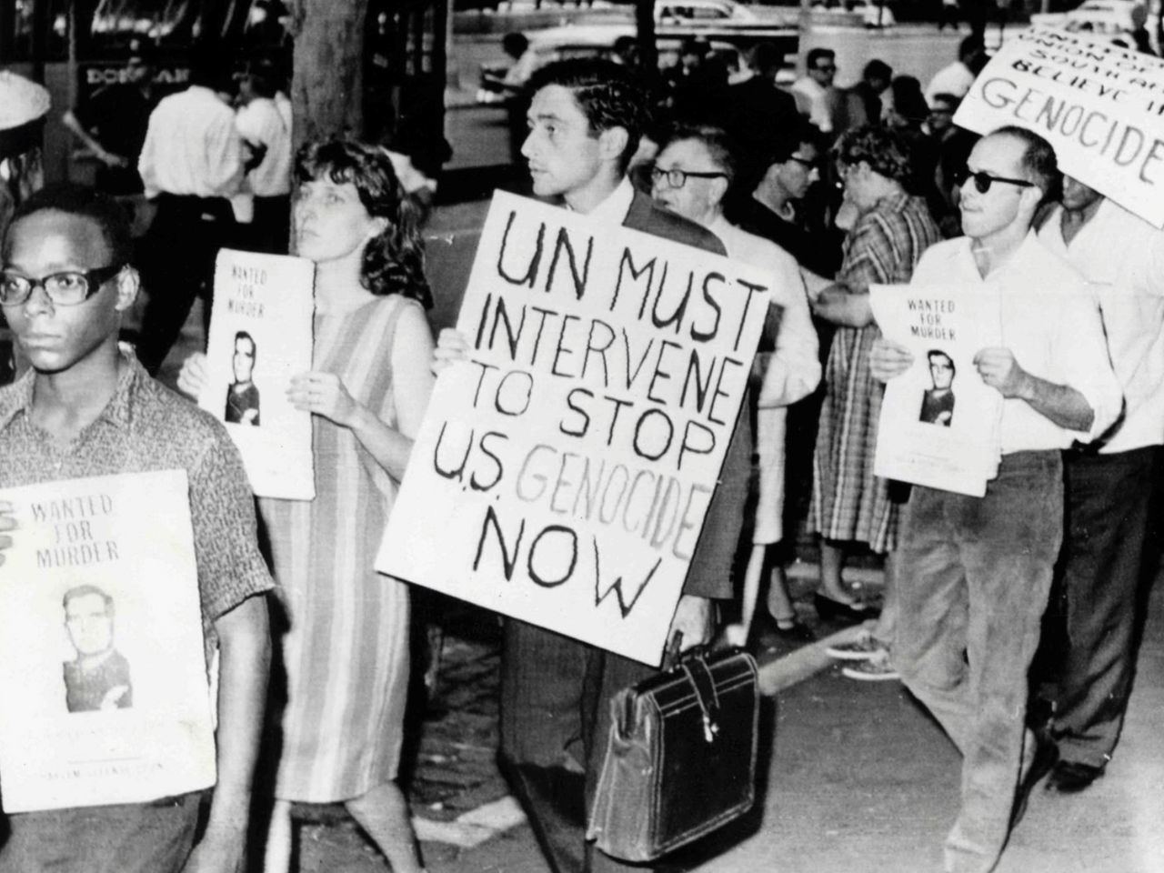 Black and white photograph of protesters holding signs that read, "Wanted for Murder" and "UN Must Intervene to Stop U.S. Genocide Now."