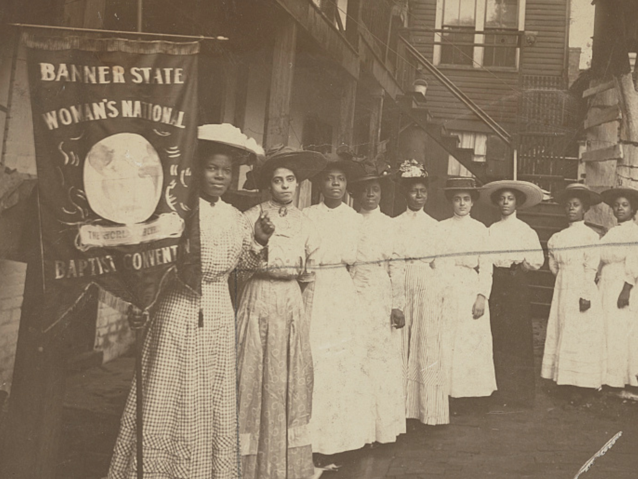 Nine Black women posed standing with one holding a banner that says "Woman's National Baptist Convention"