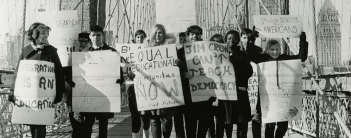A group of Black and White students carrying signs while walking across the Brooklyn Bridge; signs have slogans like "Equal Educational Opportunities Now" and "Jim Crow Can't Teach Democracy."