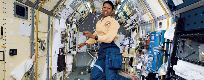 Mae Jemison, a Black woman, wearing an astronaut's uniform (yellow shirt with mission patch, blue cargo pants, grey slippers) aboard the space shuttle. She is floating in zero gravity surrounded by science stations, with a video camera floating to her left.