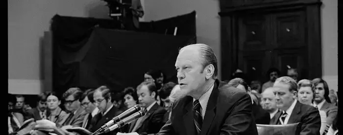 Gerald Ford seated in front of a microphone reading from a piece of paper in a House of Representatives hearing room, as others listen in among the gallery. In the background is a television camera and camera operator.