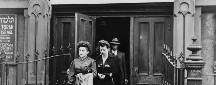 Two women in dark hats and dresses walking out of a synagogue. A man in a dark suit, white shirt, and light-colored hat is behind the women. Above the doorway of the synagogue is a sign saying "This synagogue will be open for 24 hours for special services on D-Day: All Are Welcome"