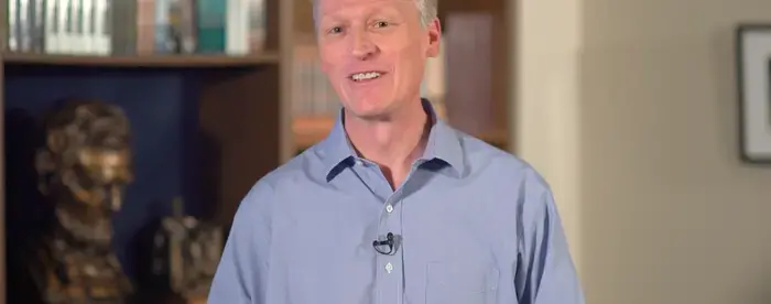 a white man wearing a blue shirt smiling while talking. In the background are books and a bust of Abraham Lincoln.