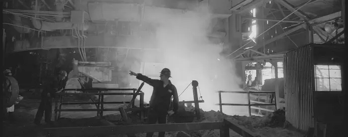 Black and white photograph of a steelworker pointing to another, with steam rising behind him. 