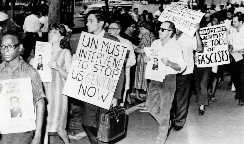 Black and white photograph of protesters holding signs that read, "Wanted for Murder" and "UN Must Intervene to Stop U.S. Genocide Now."
