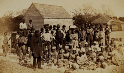 Group of emancipated enslaved people sitting in yard. With buildings visible in background.