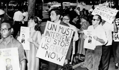 Black and white photograph of protesters holding signs that read, "Wanted for Murder" and "UN Must Intervene to Stop U.S. Genocide Now."