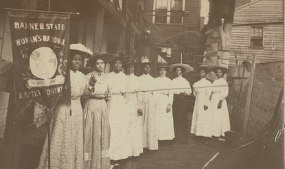 Nine Black women posed standing with one holding a banner that says "Woman's National Baptist Convention"