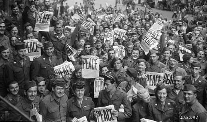 Crowd of military Service members holding up newspapers with "PEACE" headline in large letters