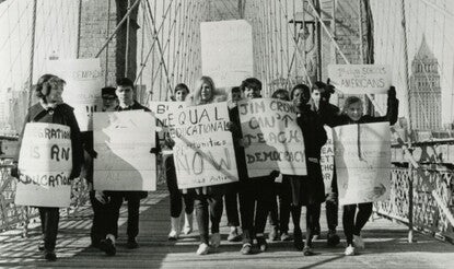 A group of Black and White students carrying signs while walking across the Brooklyn Bridge; signs have slogans like "Equal Educational Opportunities Now" and "Jim Crow Can't Teach Democracy."