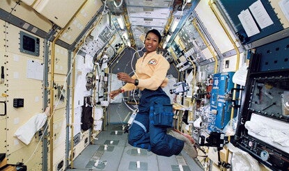 Mae Jemison, a Black woman, wearing an astronaut's uniform (yellow shirt with mission patch, blue cargo pants, grey slippers) aboard the space shuttle. She is floating in zero gravity surrounded by science stations, with a video camera floating to her left.