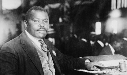 A black-and-white photograph of Marcus Garvey, a Black man wearing a dark suit, light-colored vest, white shirt, and dark tie, sitting in a wooden chair next to a desk filled with papers, holding a handkerchief in his right hand and a gavel in his left.