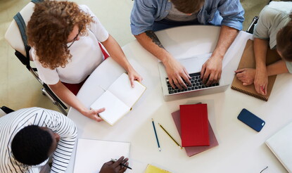A group of teens in casual wear sitting around table carrying out homework.