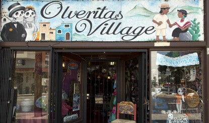 Photo shows the shop called Olveritas Village, with a "Da̕ de los Muertos" (Day of the Dead) window sign, Catrina painted on a chair back, and calaveras visible through the window and above the store. The street is part of the El Pueblo de Los Angeles Historic Monument, and referred to by Latinos as "La Placita Olvera" for a prominent local judge.