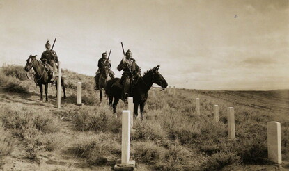 Hillside with cemetery and three Native American men on horseback.