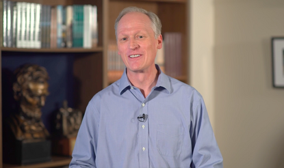 a white man wearing a blue shirt smiling while talking. In the background are books and a bust of Abraham Lincoln.