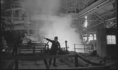Black and white photograph of a steelworker pointing to another, with steam rising behind him. 