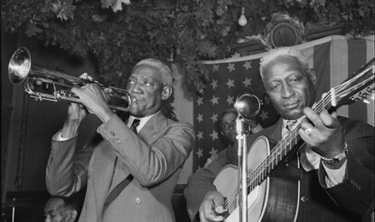 Musicians playing a guitar and the trumpet perform in front of an American flag